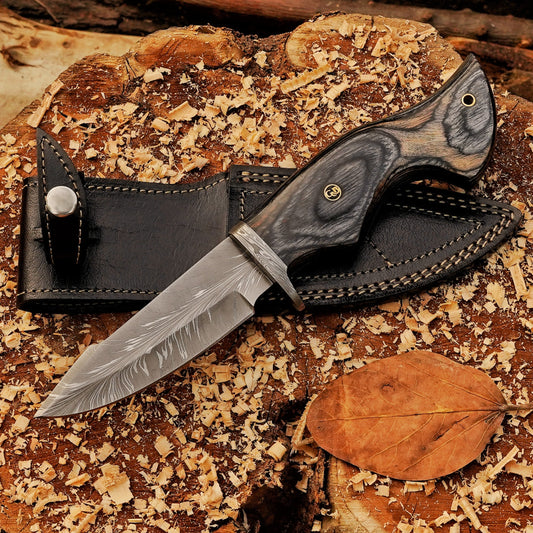 Audie Hunter Damascus hunting knife displayed diagonally on a wood stump beside its black leather sheath.
